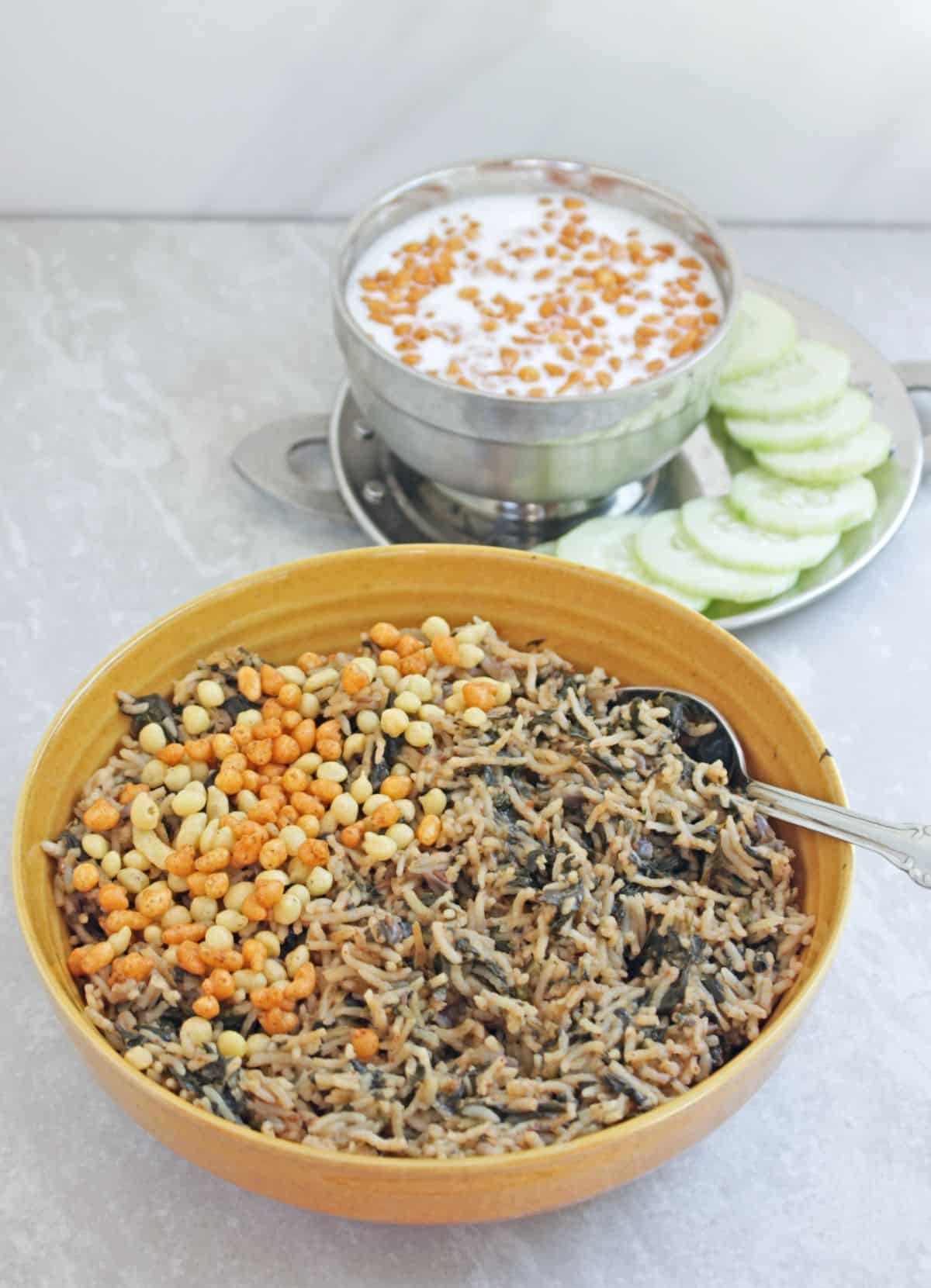 Spinach rice in a yellow bowl with boondi sprinkled on top and with raita and cucumber on side.