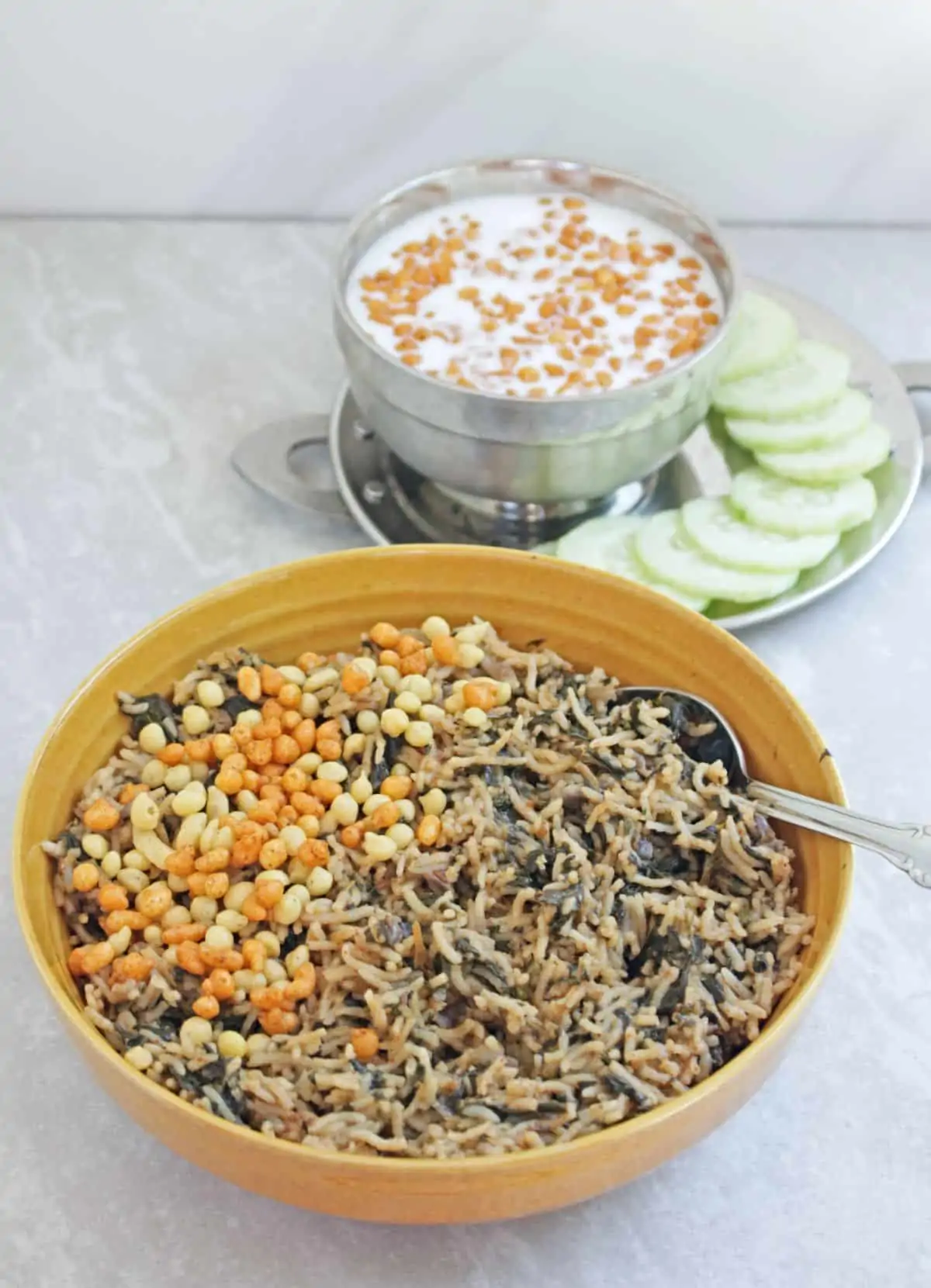 Spinach rice in a yellow bowl with boondi sprinkled on top and with raita and cucumber on side. 