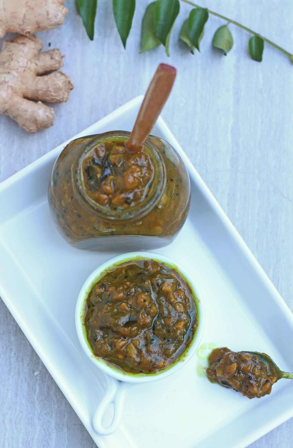 puli inji pickle in a glass jar and white bowl with ginger and curry leaves in the background